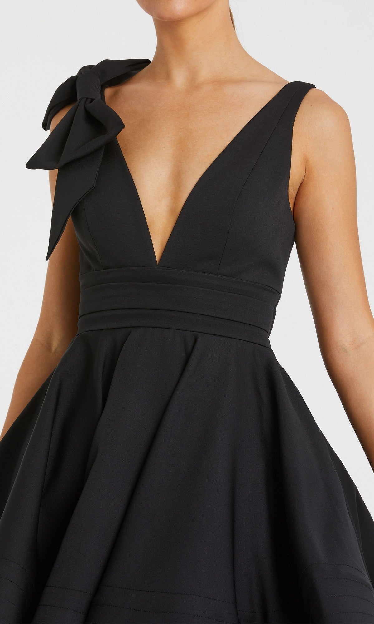 A woman wears the Mac Duggal 49971 short wedding guest dress—a sleeveless black design with a deep V-neckline, fitted waist, A-line skirt, and a statement bow on one shoulder.